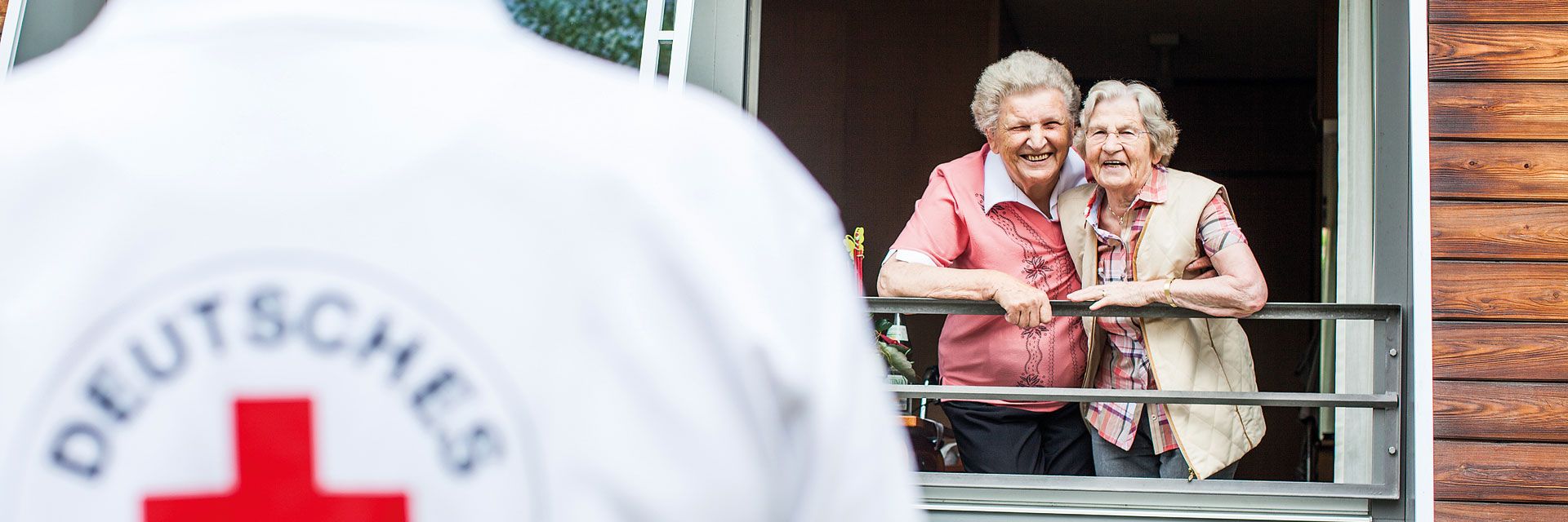 Foto: Zwei ältere Damen lehnen sich aus einem großen Fenster und blicken freudig den nahenden DRK-Mitarbeiter an.