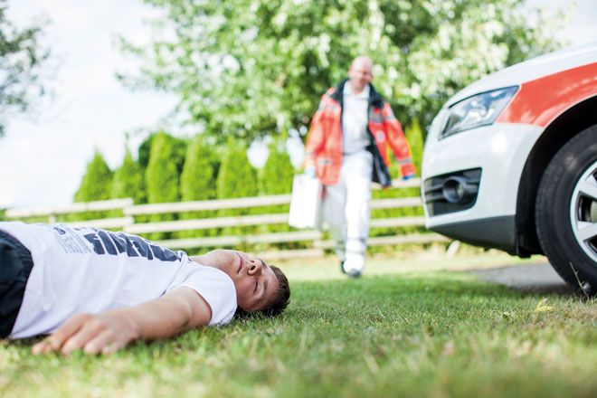 Foto: Ein junger Mann liegt bewusstlos auf einer Wiese. Ein DRK-Mitarbeiter mit Koffer nähert sich dem Patienten. Am Rande des Fotos ist ein Fahrzeug des Deutschen Roten Kreuzes zu sehen.